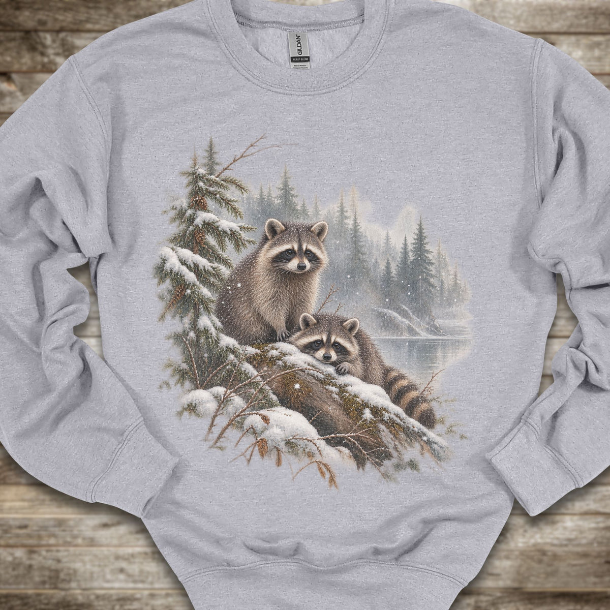 A light gray crewneck sweatshirt featuring a detailed graphic print of two raccoons in a snowy pine forest. The sweatshirt is laid flat on a rustic wooden surface.