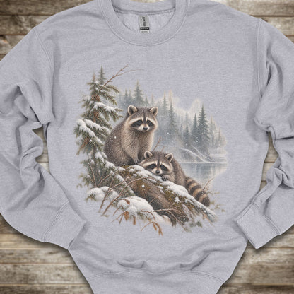 A light gray crewneck sweatshirt featuring a detailed graphic print of two raccoons in a snowy pine forest. The sweatshirt is laid flat on a rustic wooden surface.