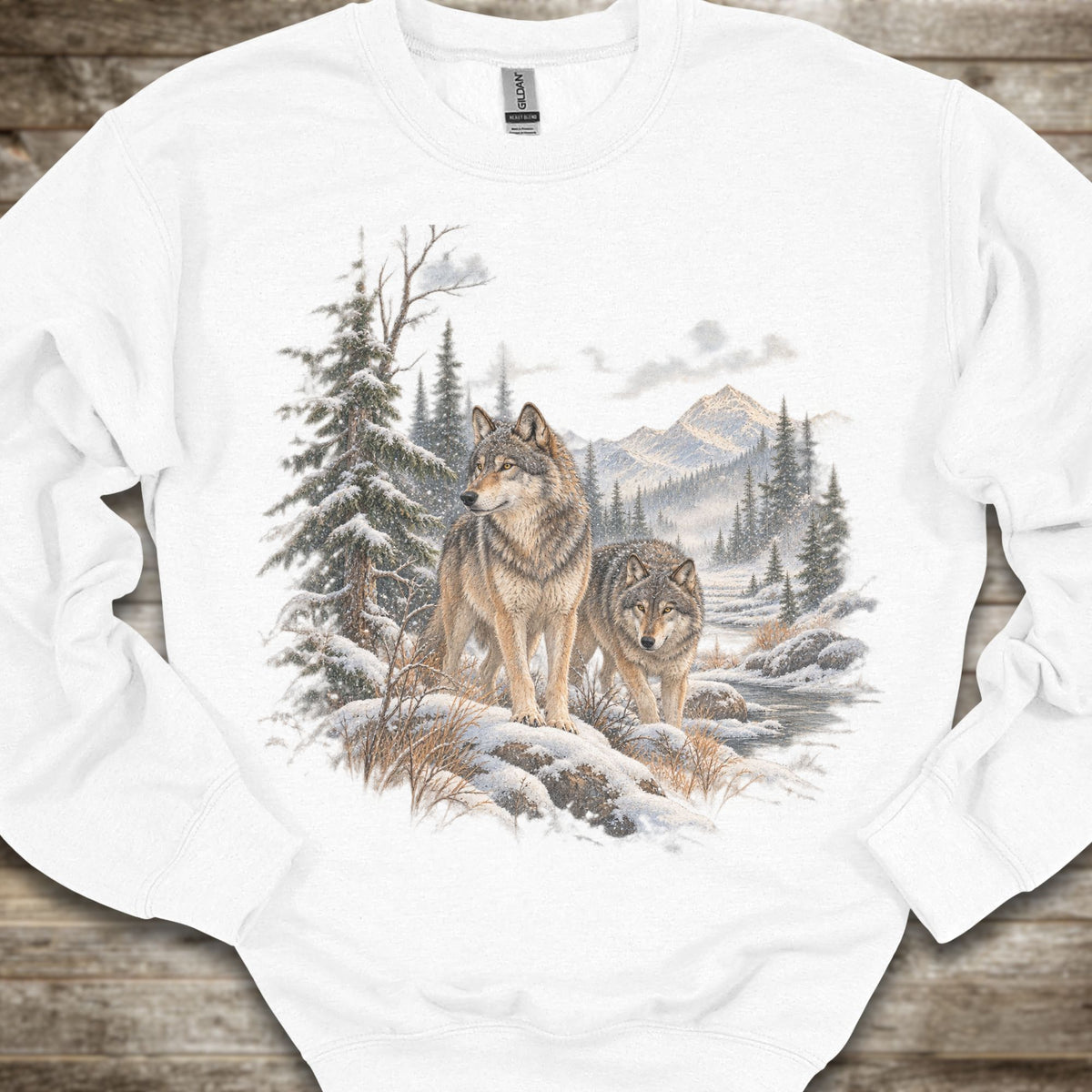 A white crewneck sweatshirt featuring a detailed graphic print of two wolves in a snowy mountain forest, laid flat on a rustic wooden surface.