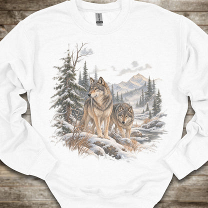 A white crewneck sweatshirt featuring a detailed graphic print of two wolves in a snowy mountain forest, laid flat on a rustic wooden surface.