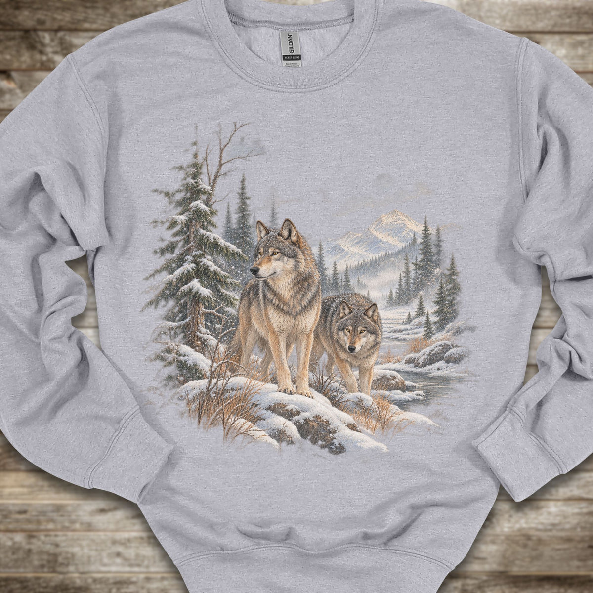 A light gray crewneck sweatshirt featuring a detailed graphic print of two wolves in a snowy winter forest, laid flat on a rustic wooden surface.