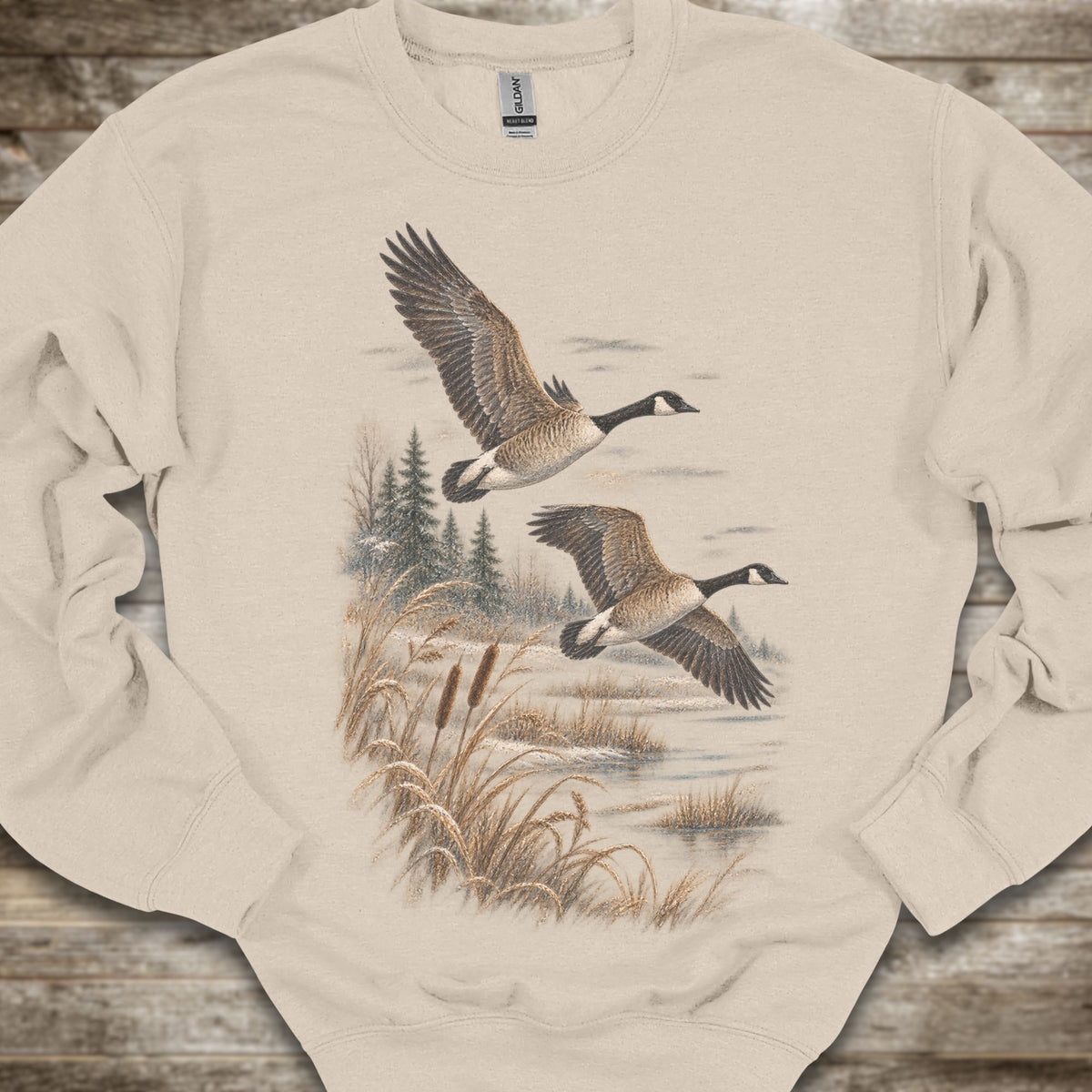 A beige crewneck sweatshirt featuring a graphic print of two Canada geese flying over a marshy landscape with reeds and pine trees, laid flat on a rustic wooden background.