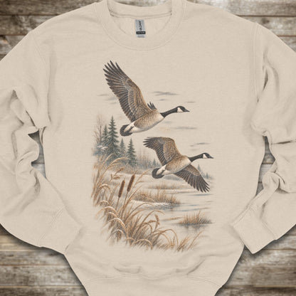 A beige crewneck sweatshirt featuring a graphic print of two Canada geese flying over a marshy landscape with reeds and pine trees, laid flat on a rustic wooden background.