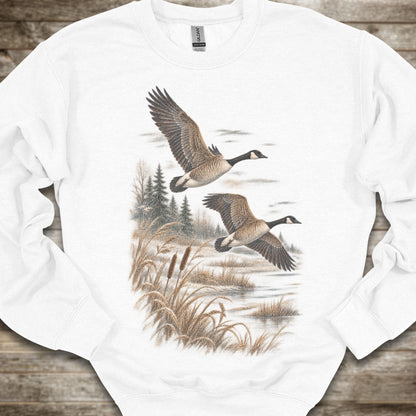 A white long-sleeve crewneck sweatshirt featuring a detailed graphic print of two Canada geese flying over a marshy landscape with reeds and pine trees, laid flat on a rustic wooden surface.
