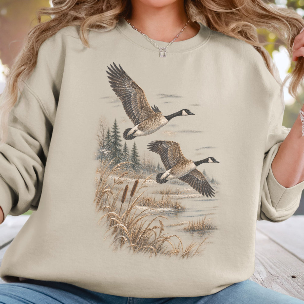 A woman wearing a beige crewneck sweatshirt with a detailed graphic print of two flying Canada geese in a marshy landscape. The sweatshirt is shown on a model in an outdoor setting.