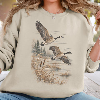 A woman wearing a beige crewneck sweatshirt with a detailed graphic print of two flying Canada geese in a marshy landscape. The sweatshirt is shown on a model in an outdoor setting.
