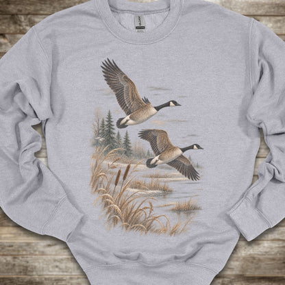 A sport grey long-sleeve crewneck sweatshirt featuring a detailed graphic print of two Canada geese flying over a marshy landscape with reeds and pine trees, laid flat on a rustic wooden surface.