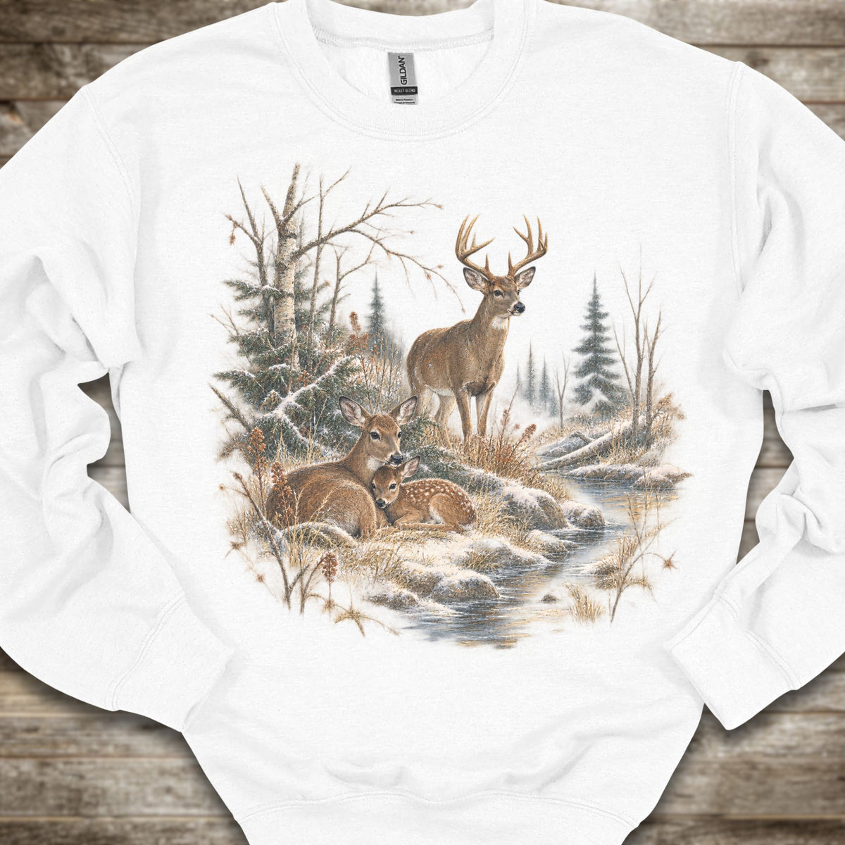 A white crewneck sweatshirt featuring a detailed winter forest graphic with deer, trees, and a stream, laid flat on a rustic wooden surface.