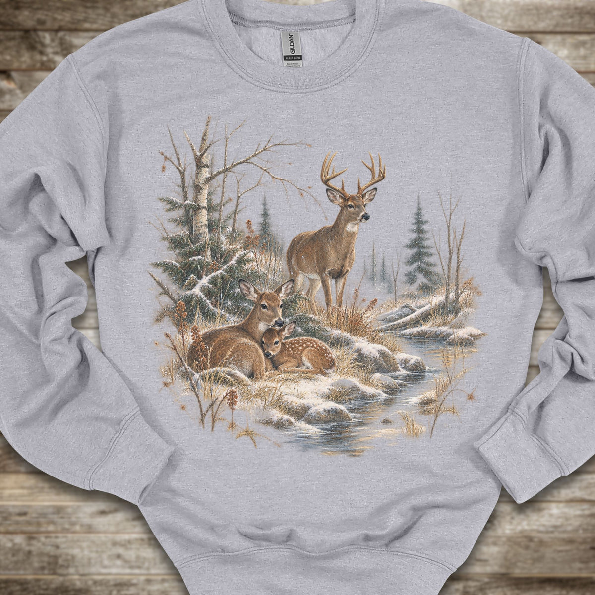 A light gray crewneck sweatshirt featuring a detailed graphic print of a winter forest scene with deer, trees, and a stream, laid flat on a rustic wooden surface.