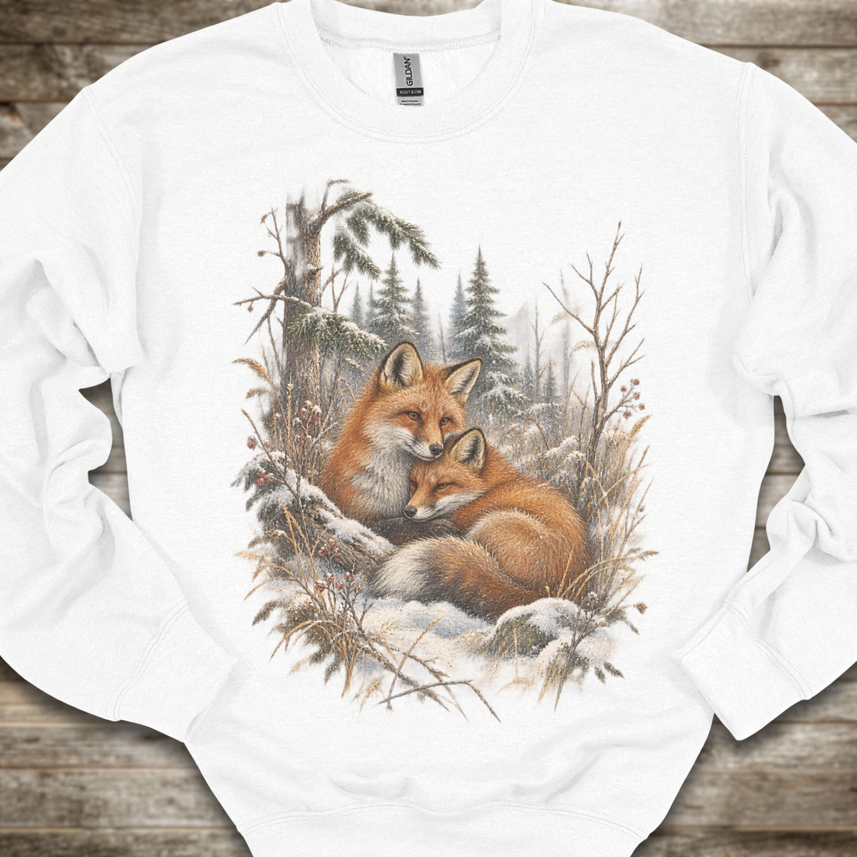 A white long-sleeve crewneck sweatshirt featuring a detailed graphic print of two red foxes resting in a snowy winter forest. The sweatshirt is laid flat on a rustic wooden surface.