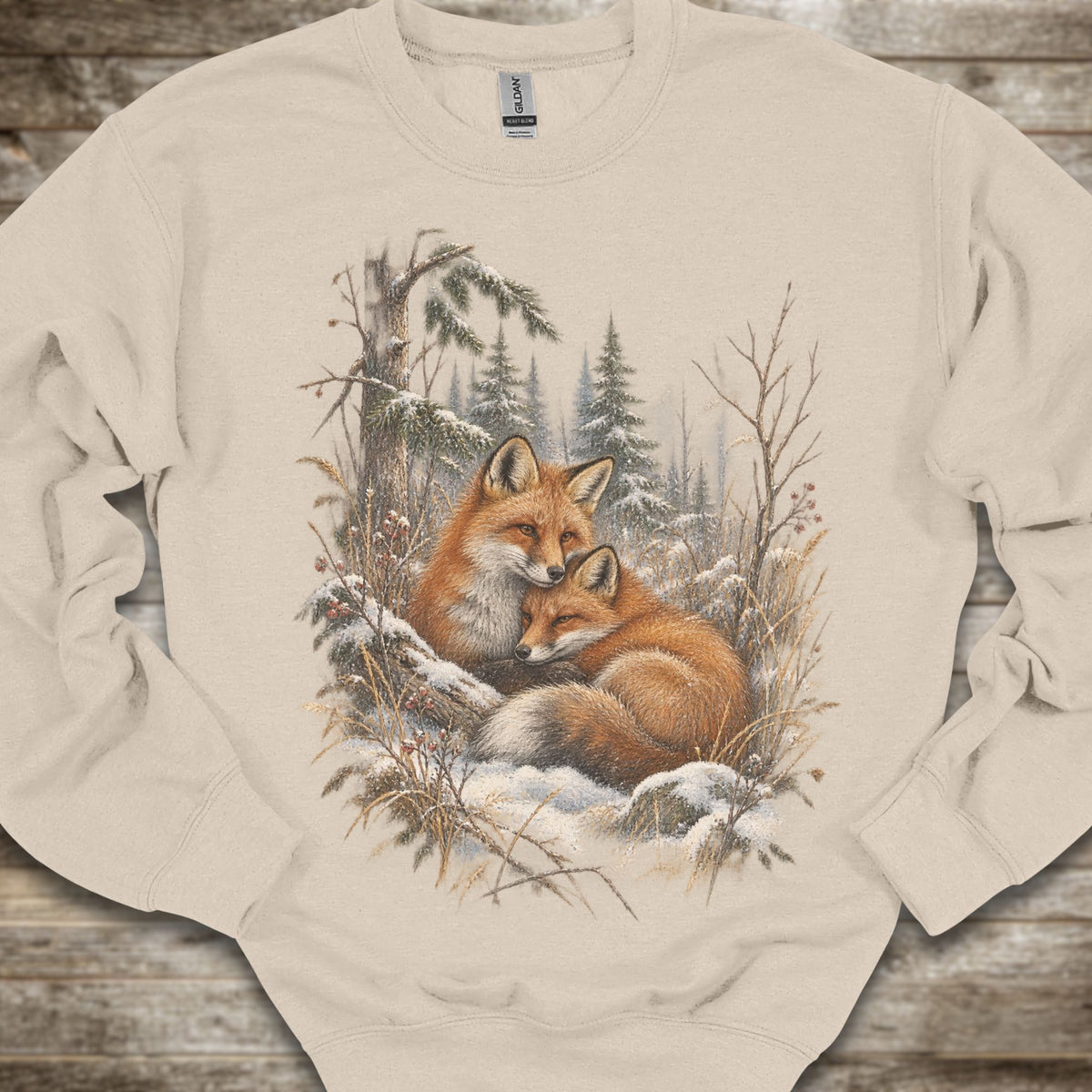 A beige crewneck sweatshirt featuring a detailed graphic print of two foxes resting in a snowy winter forest, laid flat on a rustic wooden surface.
