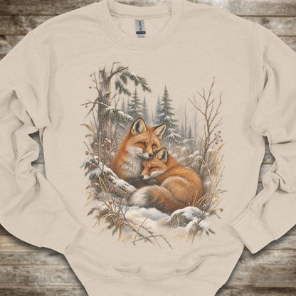 A beige crewneck sweatshirt featuring a detailed graphic print of two foxes resting in a snowy winter forest, laid flat on a rustic wooden surface.