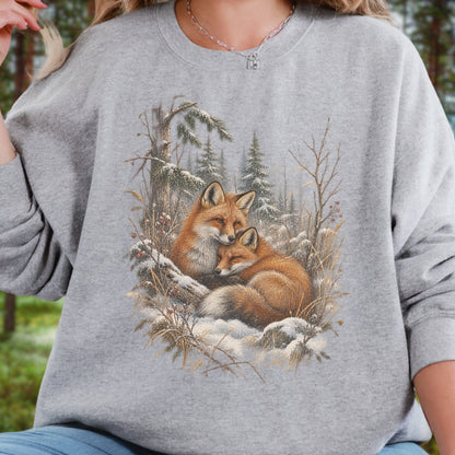 A person wearing a heather gray crewneck sweatshirt with a detailed graphic print of two foxes resting in a snowy forest. The background is a blurred outdoor scene.