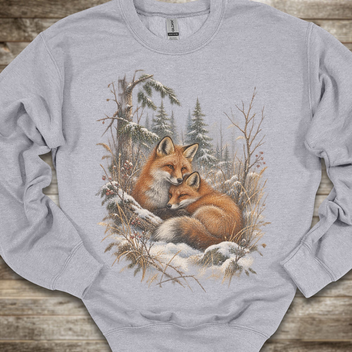 A light gray crewneck sweatshirt featuring a detailed graphic print of two red foxes resting in a snowy winter forest, laid flat on a rustic wooden surface.