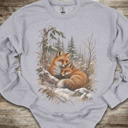 A light gray crewneck sweatshirt featuring a detailed graphic print of two red foxes resting in a snowy winter forest, laid flat on a rustic wooden surface.