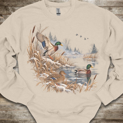 A beige crewneck sweatshirt featuring a detailed graphic print of mallards in a snowy winter landscape, laid flat on a rustic wooden background.