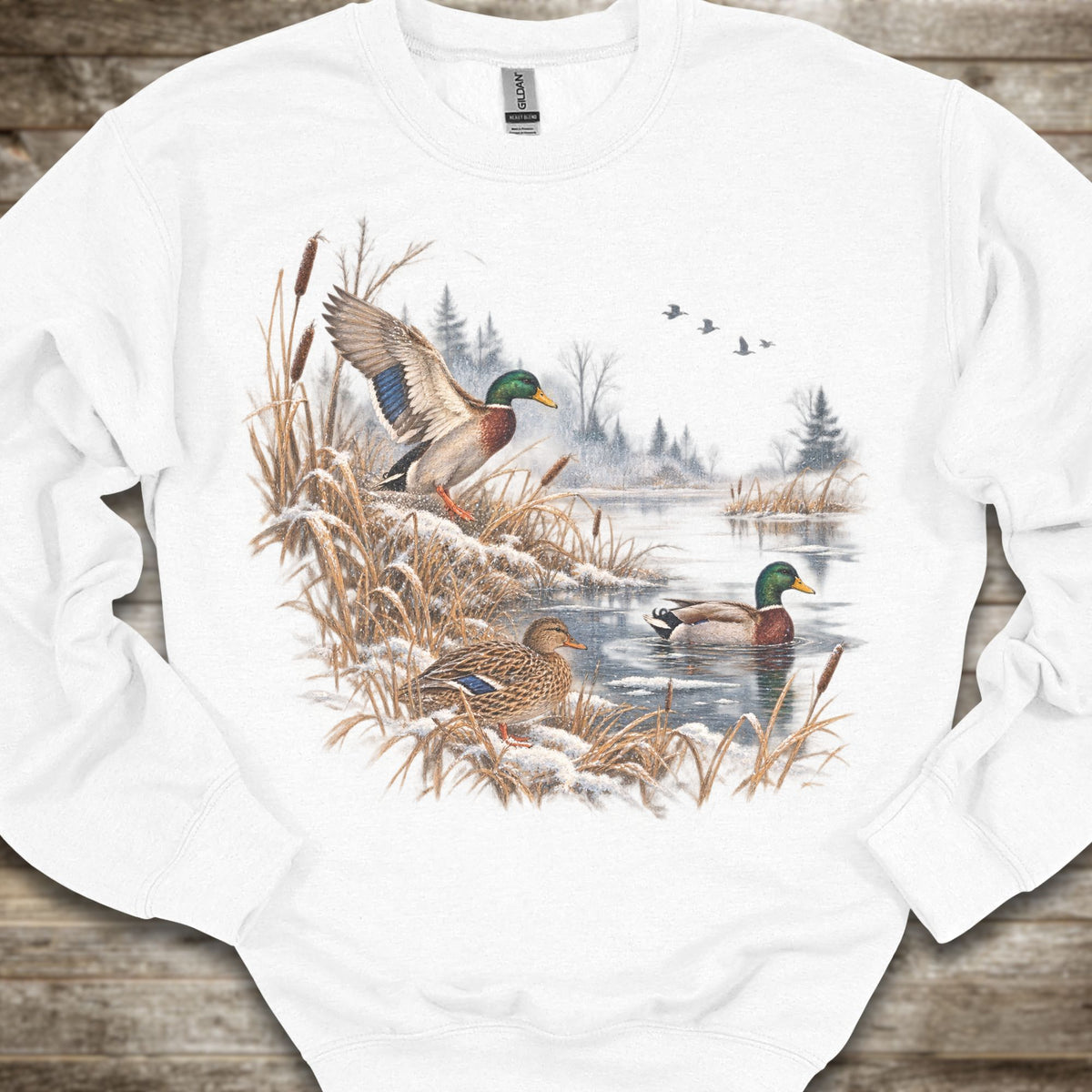 A white crewneck sweatshirt featuring a detailed graphic print of mallards in a snowy winter landscape by a pond, laid flat on a rustic wooden background.