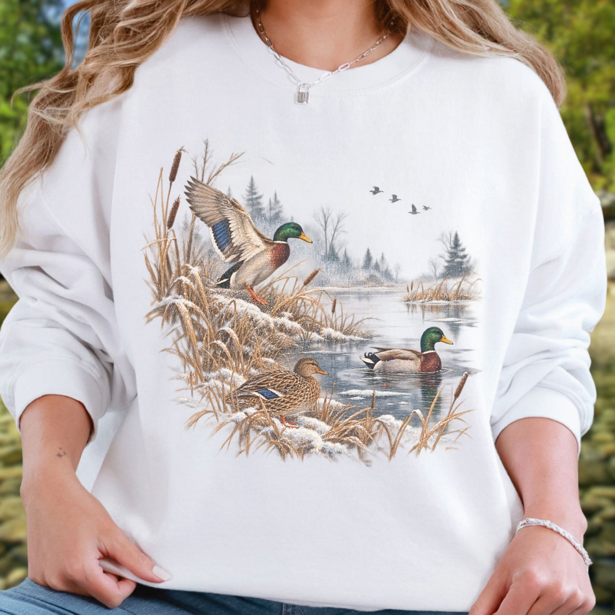 A woman wearing a white crewneck sweatshirt with a detailed graphic print of mallards by a snowy pond, with a blurred outdoor background.