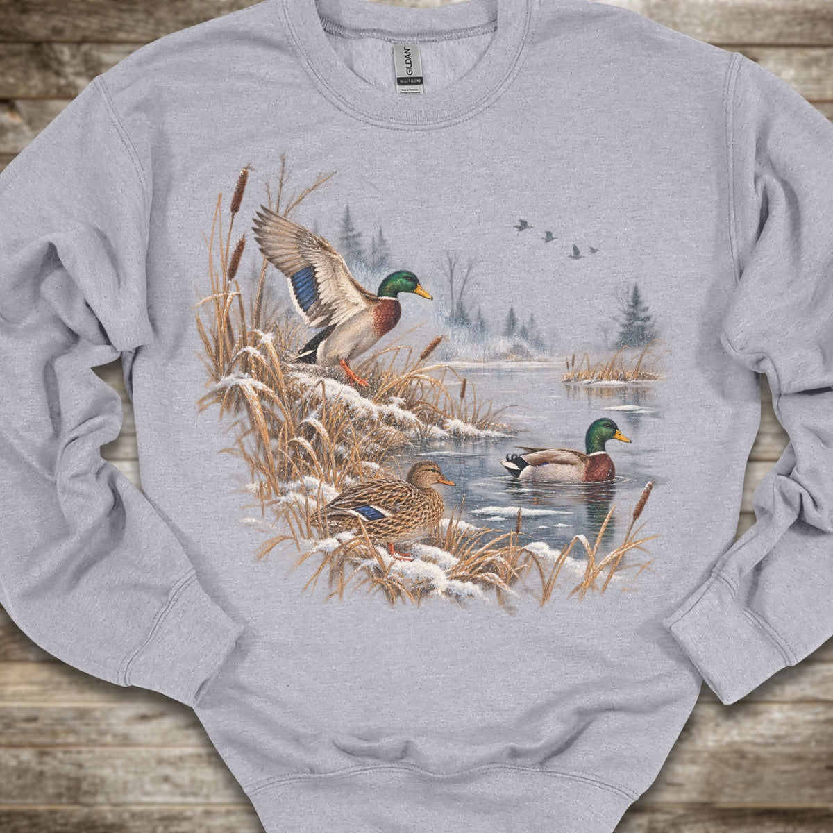 A light gray crewneck sweatshirt featuring a detailed graphic print of mallards in a snowy winter landscape, laid flat on a rustic wooden surface.