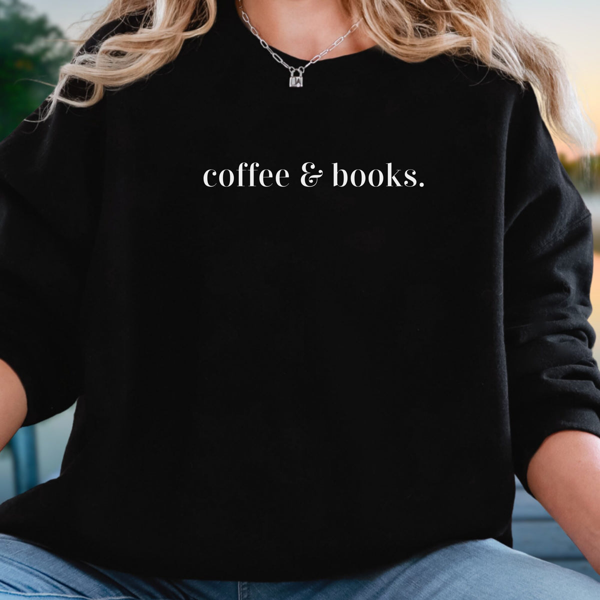 A person wearing a black long-sleeve sweatshirt with the phrase 'coffee & books.' printed in white serif text across the chest, paired with blue jeans and a silver necklace.
