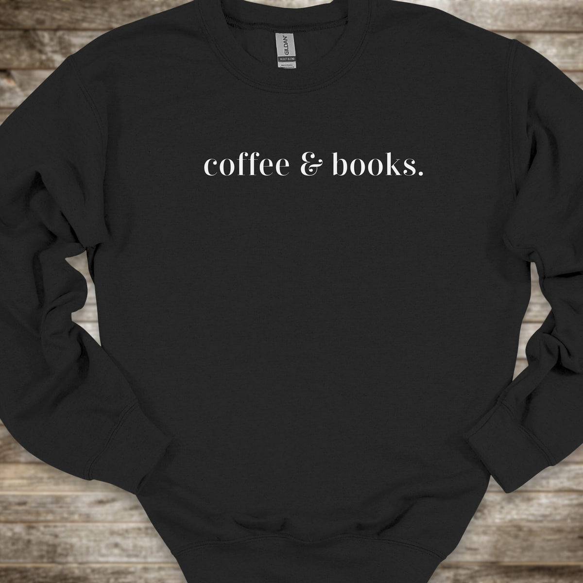 A black crewneck sweatshirt with the phrase 'coffee & books.' printed in white serif text across the chest, laid flat on a rustic wooden background.