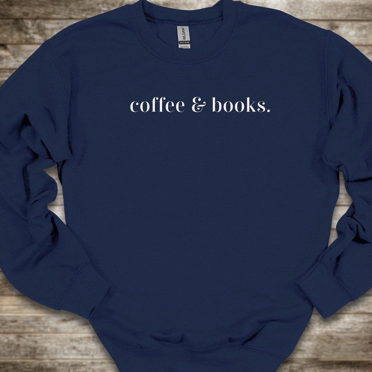 A navy blue crewneck sweatshirt with the phrase 'coffee & books.' printed in white serif text across the chest, laid flat on a rustic wooden background.