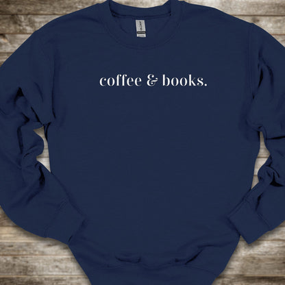A navy blue crewneck sweatshirt with the phrase 'coffee & books.' printed in white serif text across the chest, laid flat on a rustic wooden background.
