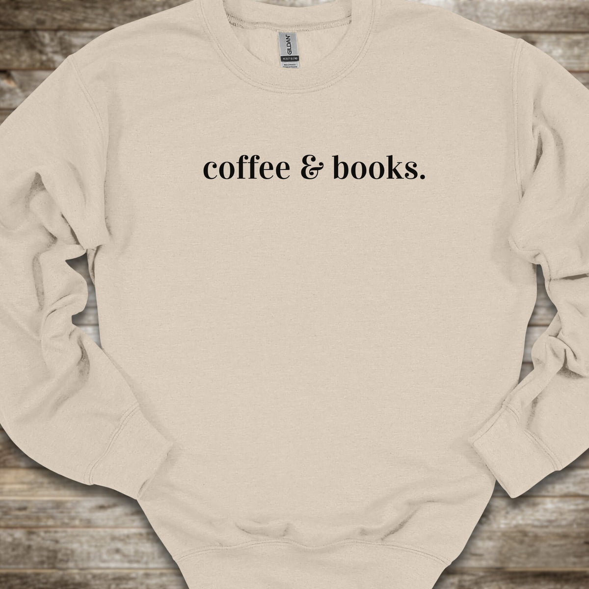 A light beige crewneck sweatshirt with the phrase 'coffee & books.' printed in black serif font across the chest, laid flat on a rustic wooden background.