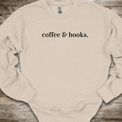 A light beige crewneck sweatshirt with the phrase 'coffee & books.' printed in black serif font across the chest, laid flat on a rustic wooden background.