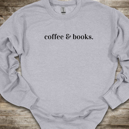 A light heather gray crewneck sweatshirt with the phrase 'coffee & books.' printed in black serif text across the chest, laid flat on a rustic wooden background.