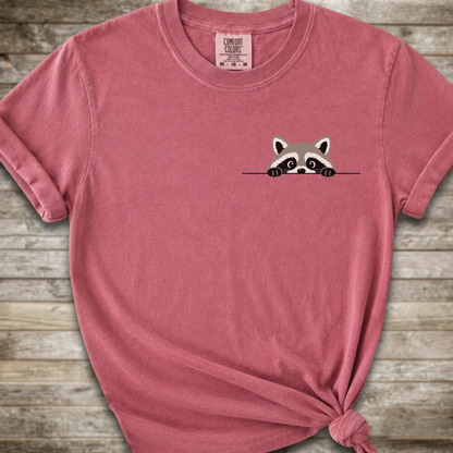 A dark pinkshort-sleeve t-shirt with a small cartoon raccoon peeking over a line on the left chest, styled with a knot at the bottom hem against a rustic wood plank background.