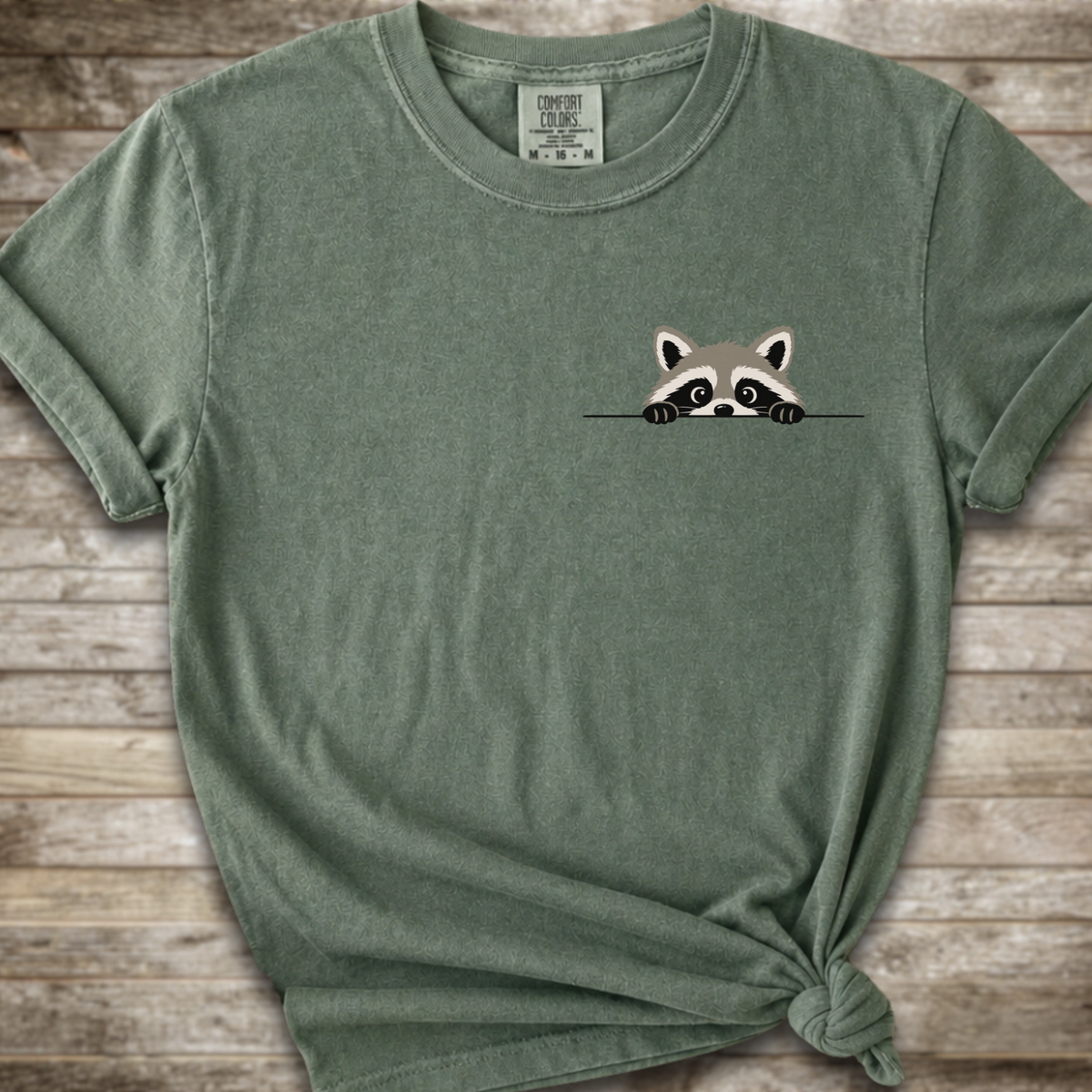 A  green heathered t-shirt with a small cartoon raccoon peeking over a line on the left chest, styled with a knot at the bottom hem on a rustic wooden background.