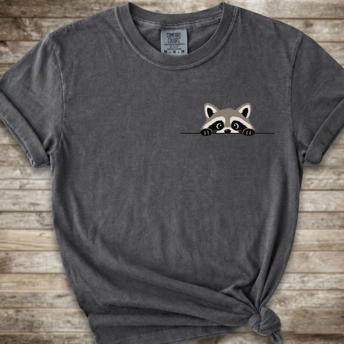 A dark gray t-shirt with a small cartoon raccoon peeking over a line on the left chest, styled with a knot at the bottom right against a rustic wooden background.