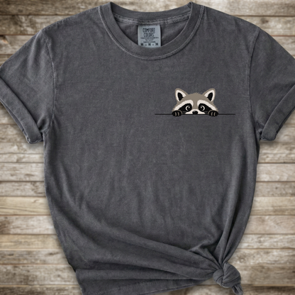 A dark gray t-shirt with a small cartoon raccoon peeking over a line on the left chest, styled with a knot at the bottom right against a rustic wooden background.