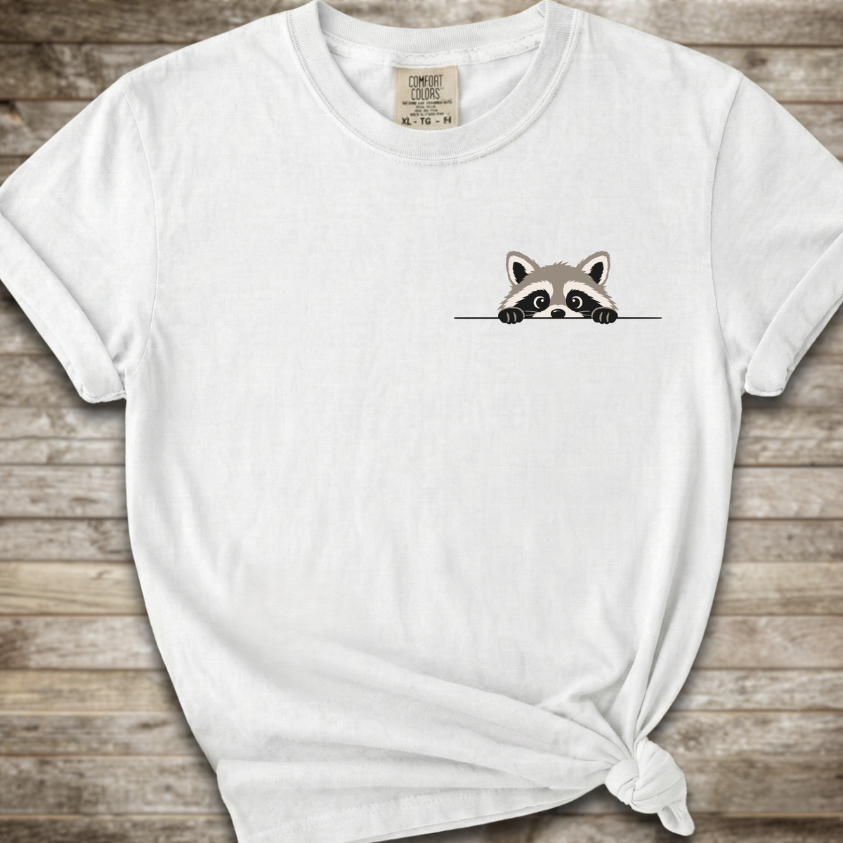 A white short-sleeve t-shirt with a small cartoon raccoon peeking over a line on the left chest, styled with a knot at the bottom right against a rustic wooden background.