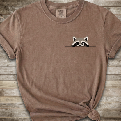 A brown short-sleeve t-shirt with a small cartoon raccoon peeking over a line on the left chest, styled with a knot at the bottom hem against a rustic wood plank background.