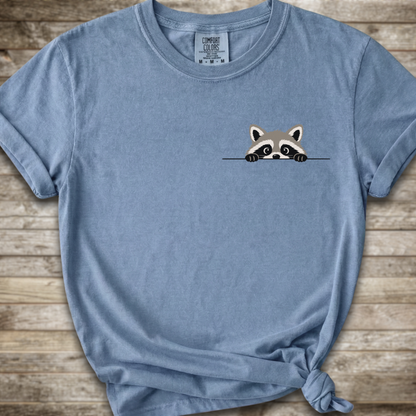 A light blue short-sleeve t-shirt with a small cartoon raccoon peeking over a line on the chest, styled with a knot at the bottom hem against a rustic wood plank background. The brand tag 'Comfort Colors' is visible inside the collar.