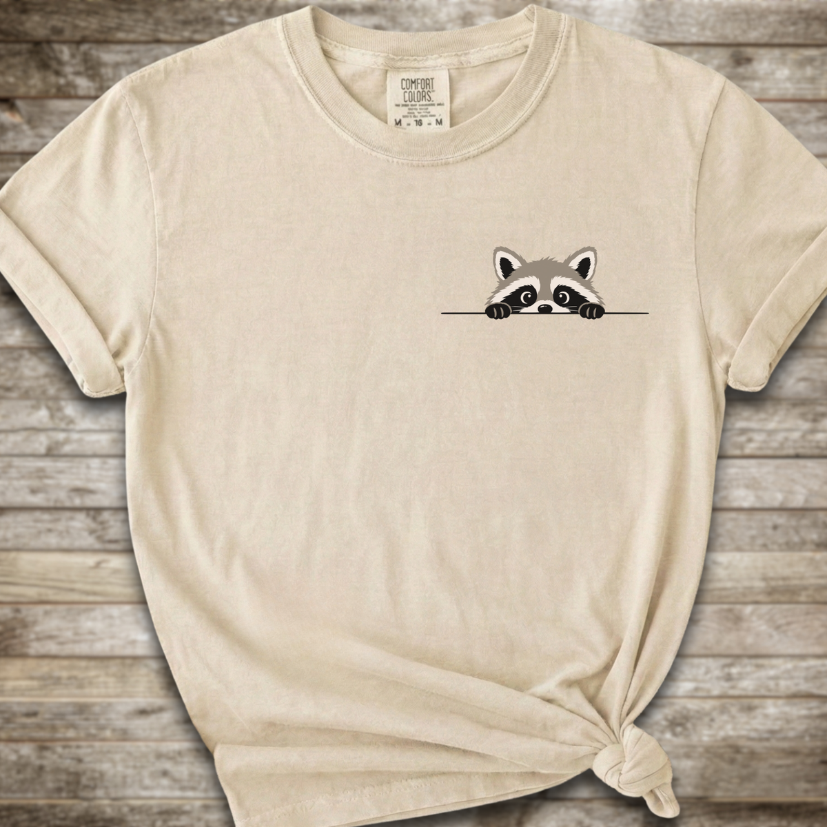 A cream-colored short-sleeve t-shirt with a small cartoon raccoon peeking over a line on the left chest, styled on a rustic wooden background with a knot at the bottom hem.
