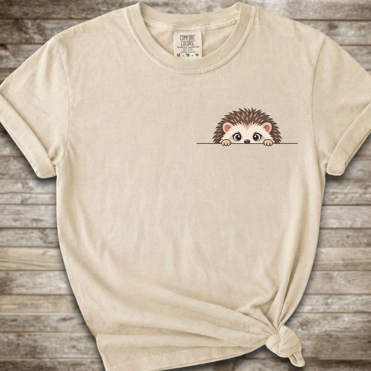 A light beige crewneck t-shirt with a small cartoon hedgehog peeking over a line on the left chest, styled on a rustic wooden background with a knot at the bottom hem.