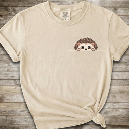 A light beige crewneck t-shirt with a small cartoon hedgehog peeking over a line on the left chest, styled on a rustic wooden background with a knot at the bottom hem.