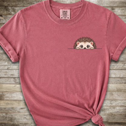 A dark pink short-sleeve t-shirt with a small cartoon hedgehog peeking out from the chest pocket, styled with a knot at the bottom hem against a rustic wood plank background.