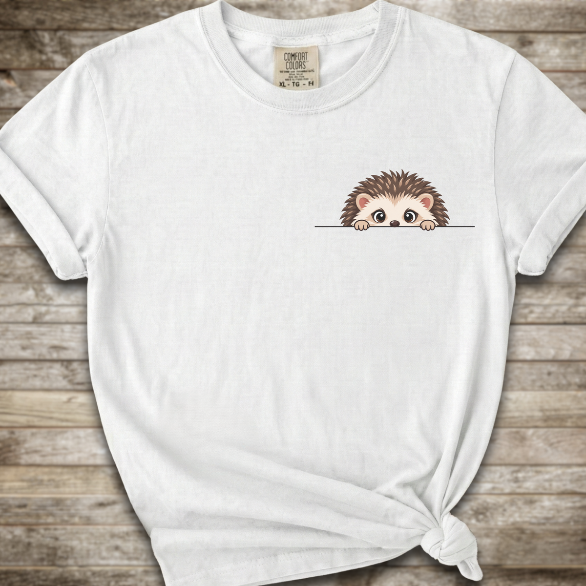 A white short-sleeve t-shirt with a small cartoon hedgehog graphic peeking over a line on the left chest, styled on a rustic wooden plank background with a knot at the bottom hem.