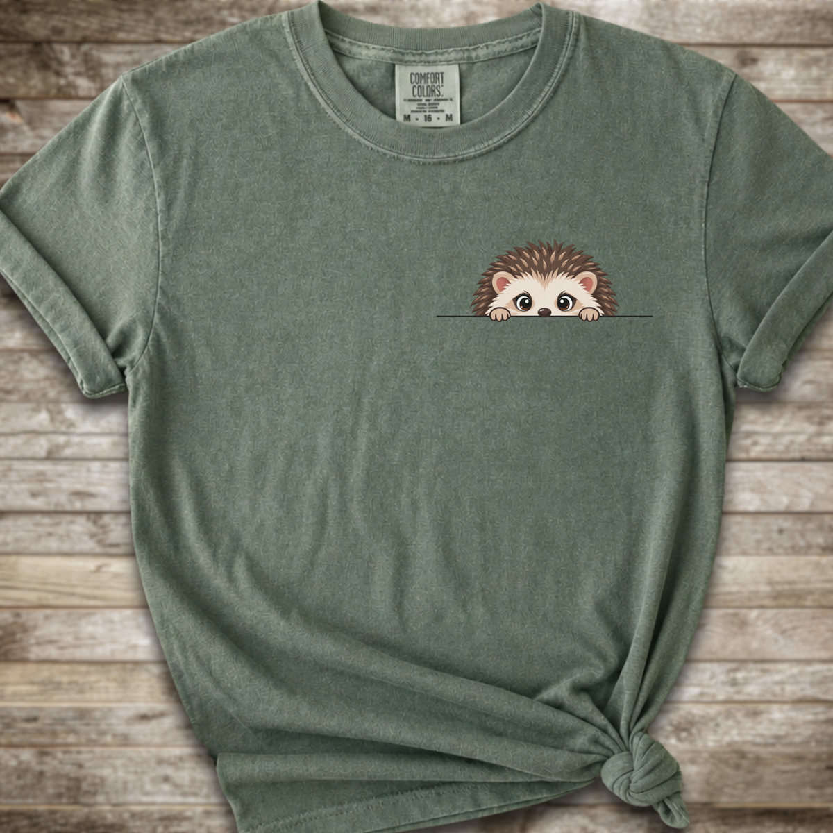 A moss green short-sleeve t-shirt with a small cartoon hedgehog peeking over a line on the left chest, styled with a knot at the bottom hem against a rustic wood plank background.