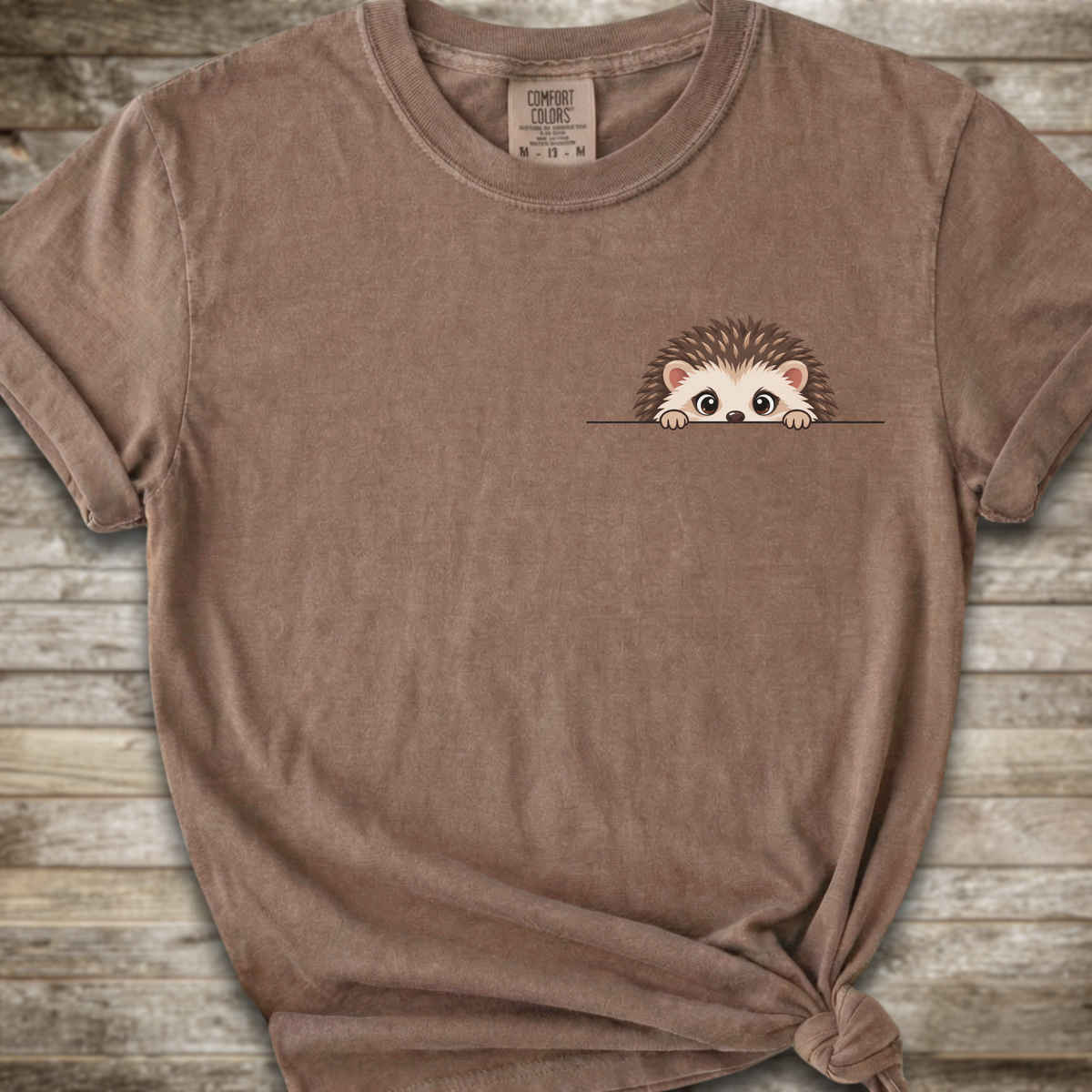 A brown short-sleeve t-shirt with a cute cartoon hedgehog peeking out from the chest pocket, styled on a rustic wooden background.