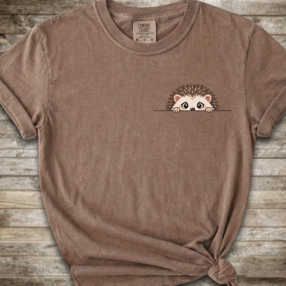 A brown short-sleeve t-shirt with a cute cartoon hedgehog peeking out from the chest pocket, styled on a rustic wooden background.
