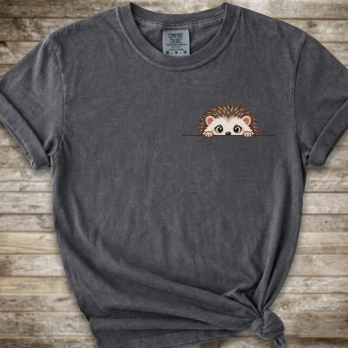 A dark gray  t-shirt with a cute cartoon hedgehog peeking out from the chest pocket, styled with a knot at the bottom hem against a rustic wooden background.