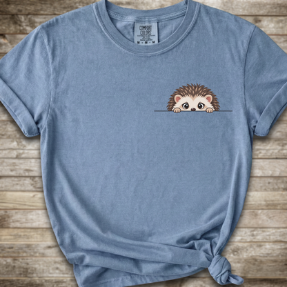 A light blue short-sleeve t-shirt with a small cartoon hedgehog peeking over a line on the chest, styled with a knot at the bottom hem against a rustic wood plank background.