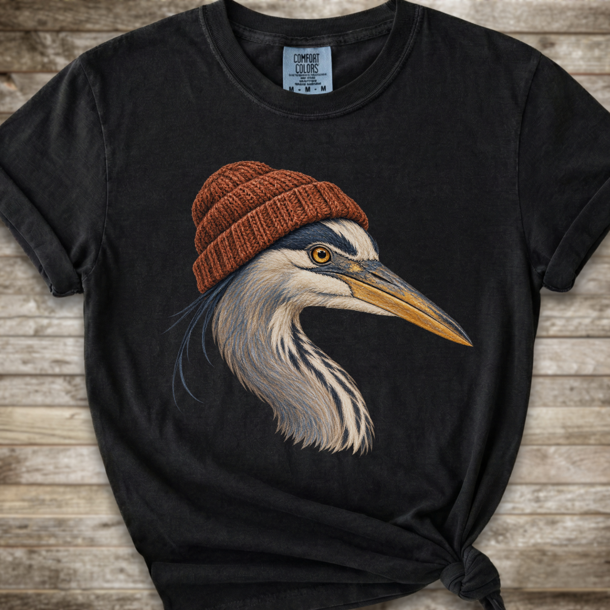 A black graphic t-shirt featuring a detailed illustration of a heron wearing a brown knit beanie, laid flat on a rustic wooden background.