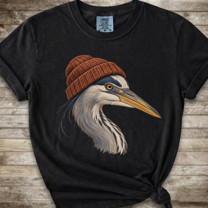 A black graphic t-shirt featuring a detailed illustration of a heron wearing a brown knit beanie, laid flat on a rustic wooden background.