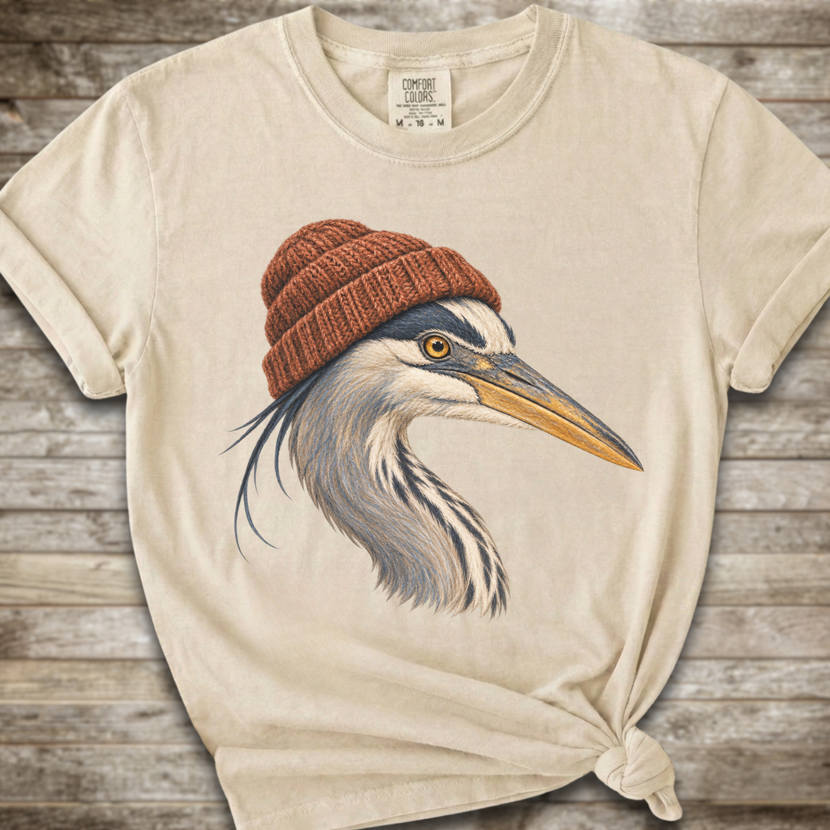 A cream-colored t-shirt with a graphic of a heron wearing a brown knit beanie, styled with a knot at the bottom right against a rustic wood background.